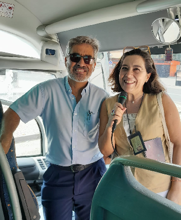 Two people smiling inside a bus, one holding a microphone.