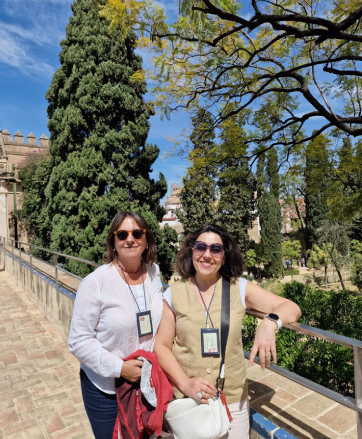 Two women posing outdoors with trees and blue sky background.