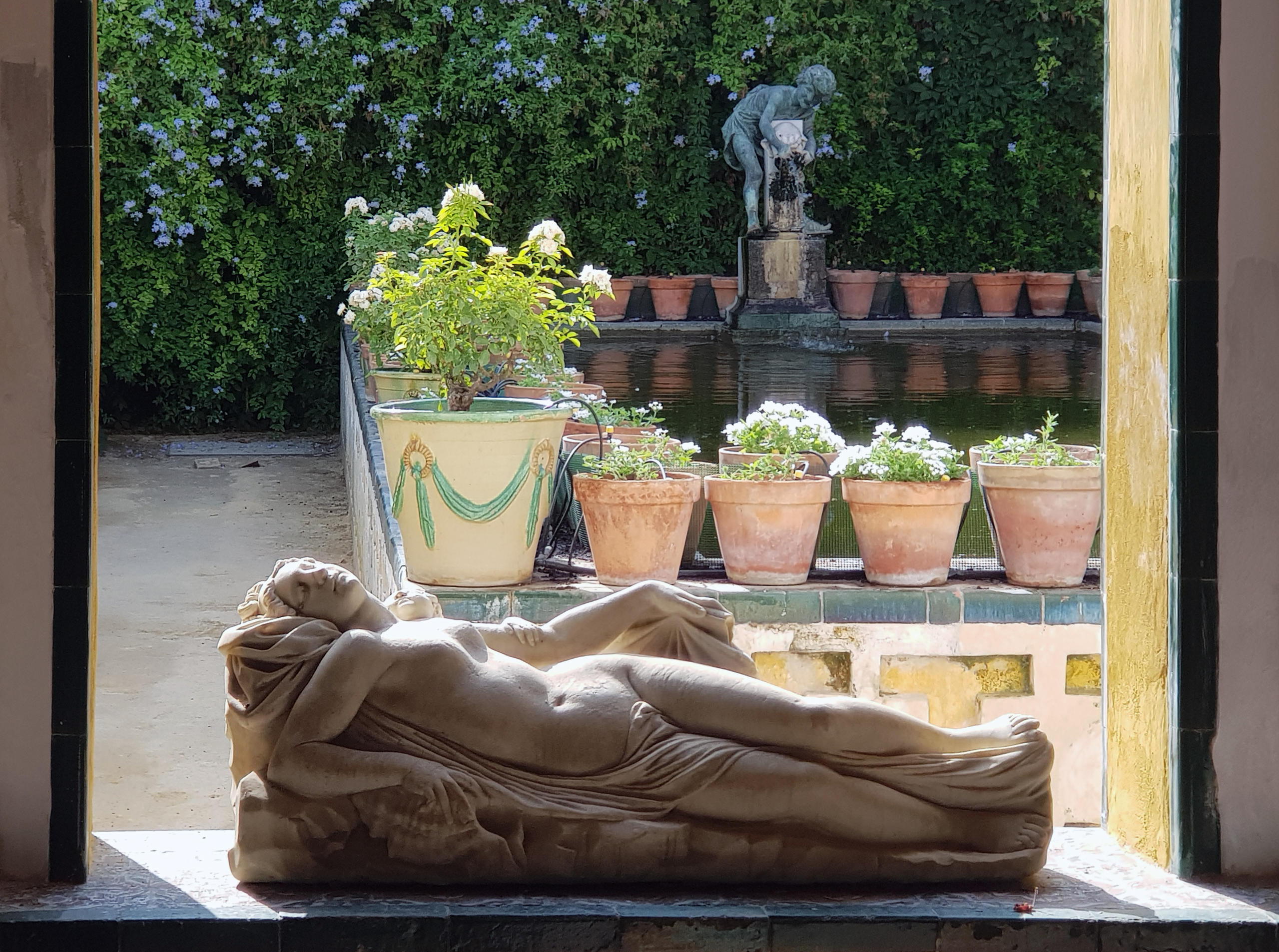 Reclining statue by window with garden view and potted plants.