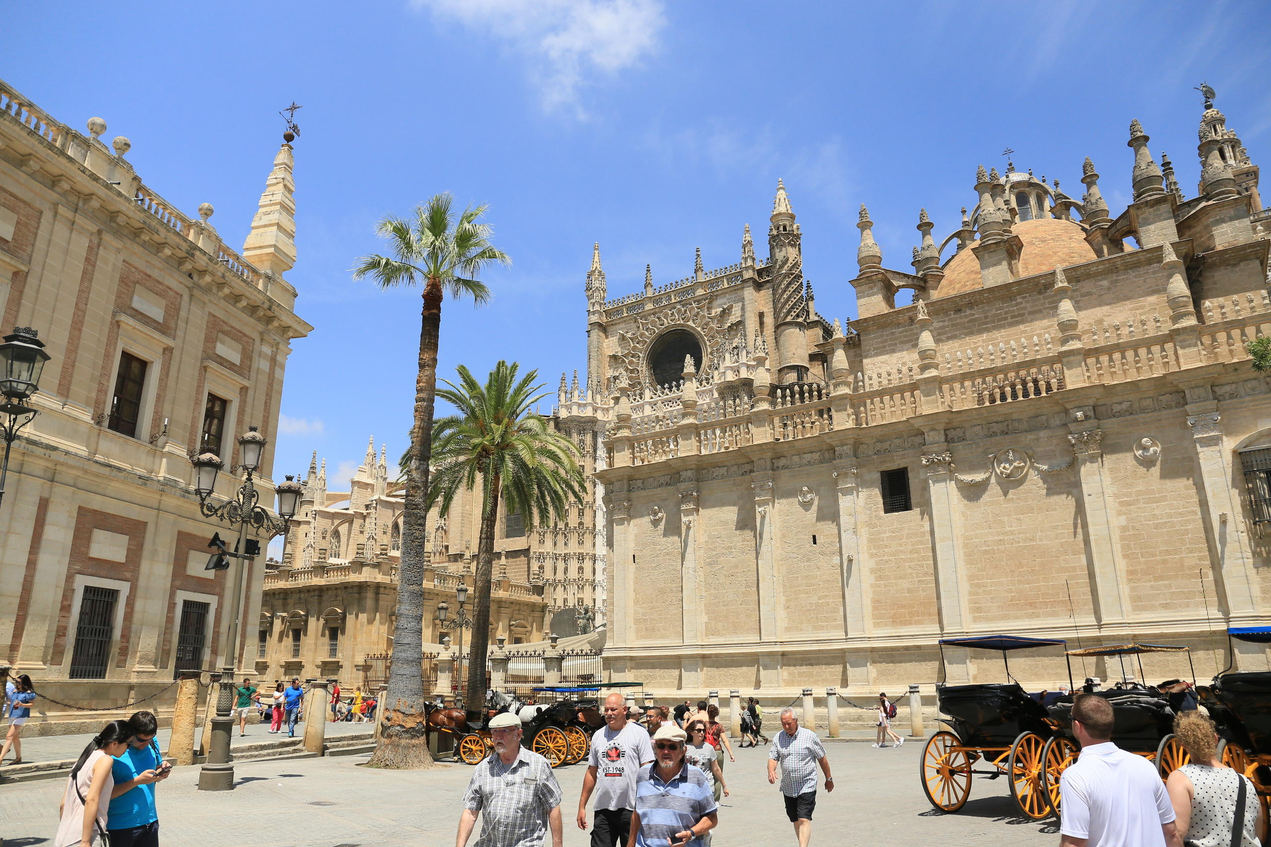 Tourists walk by historic cathedral and horse-drawn carriages under a clear blue sky.
