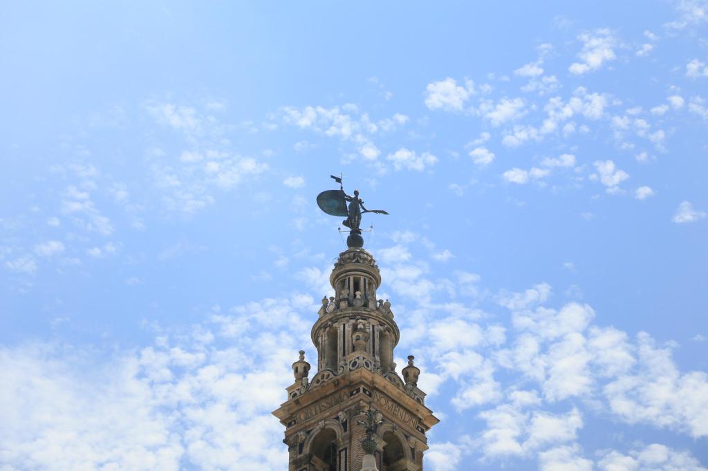Ornate tower with a weather vane against a blue sky with clouds.