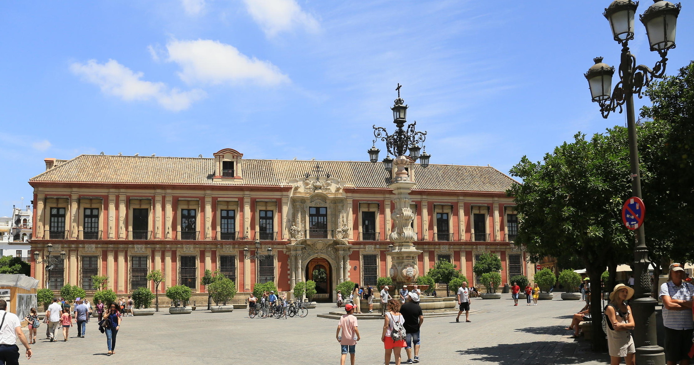 People walking near historic building with arch and ornate lantern in sunny plaza.