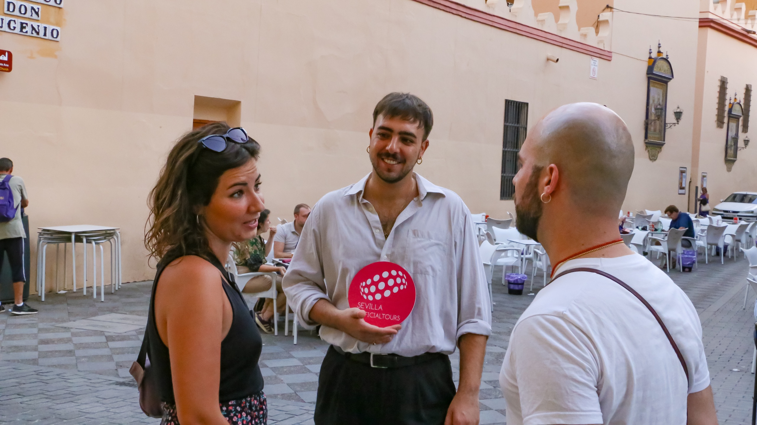 Three people talking in a courtyard with chairs and tables in the background.