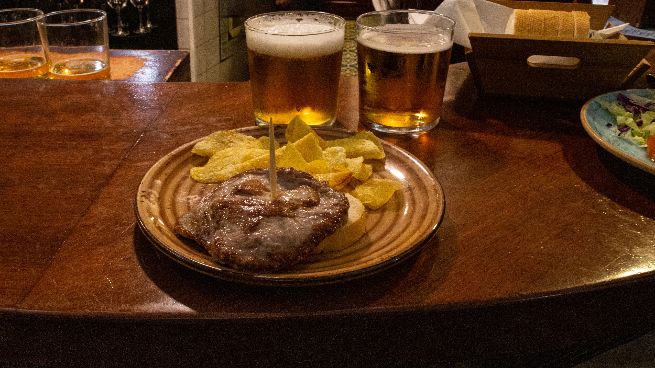 Plate with meat and chips on a wooden bar, two glasses of beer beside it.