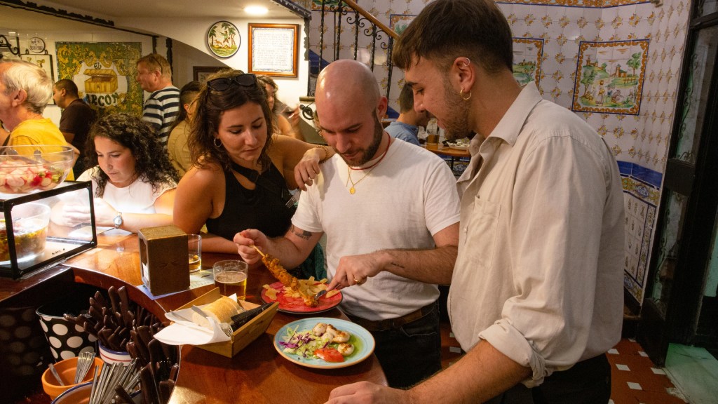Three people share food at a busy tapas bar.