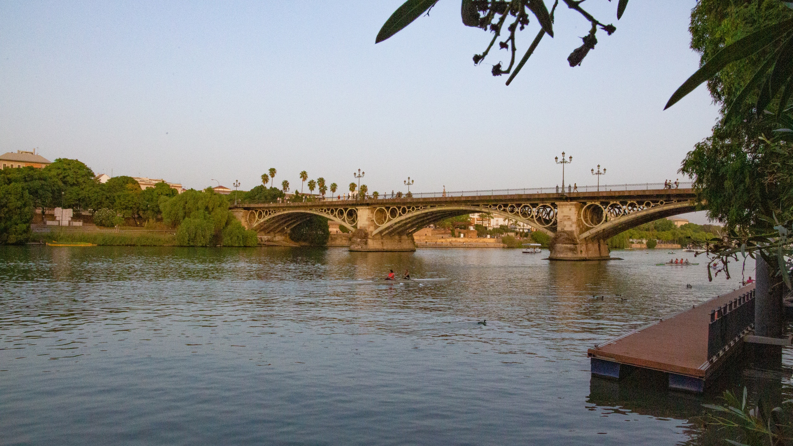 Ornate bridge over a calm river with trees and a dock at sunset.