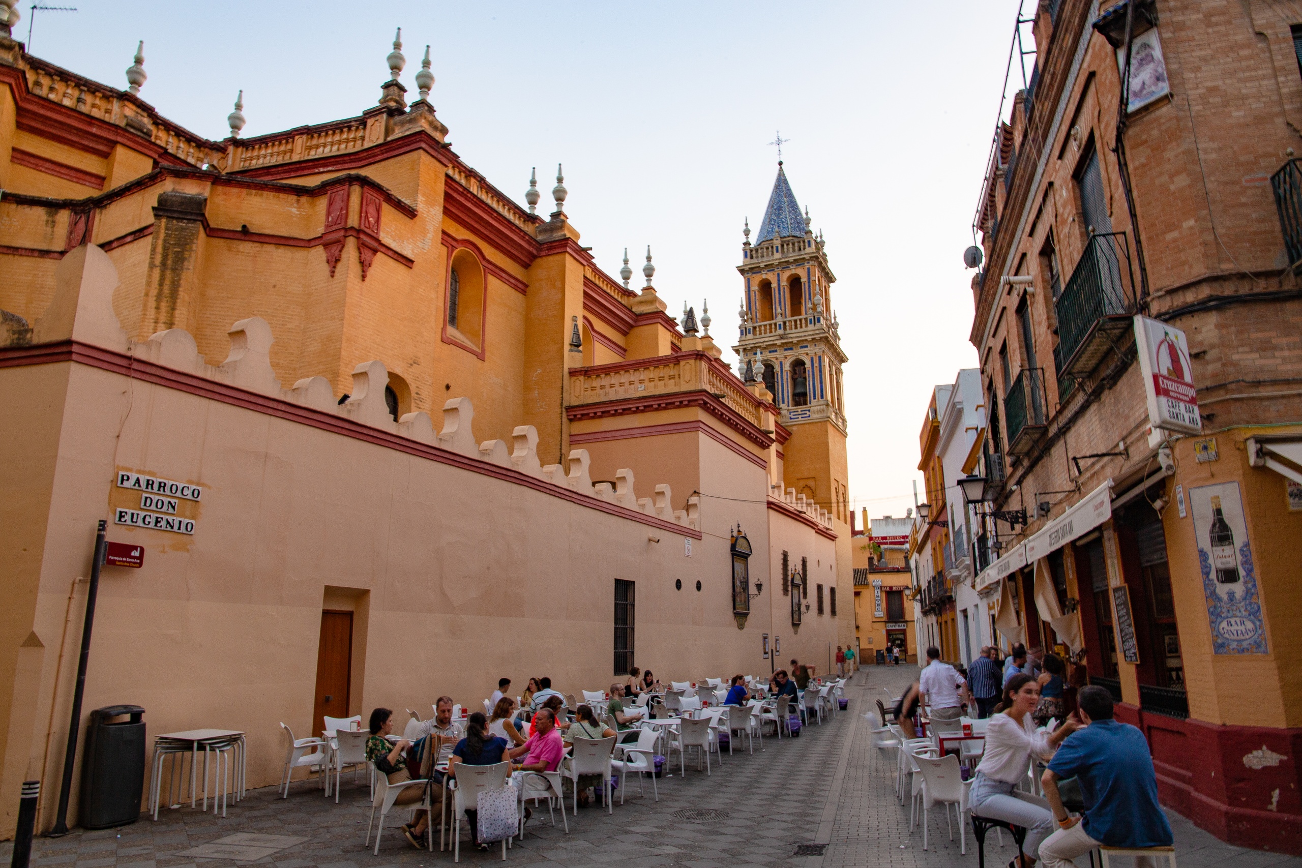 People dining on a narrow street beside an old church with a tall bell tower.