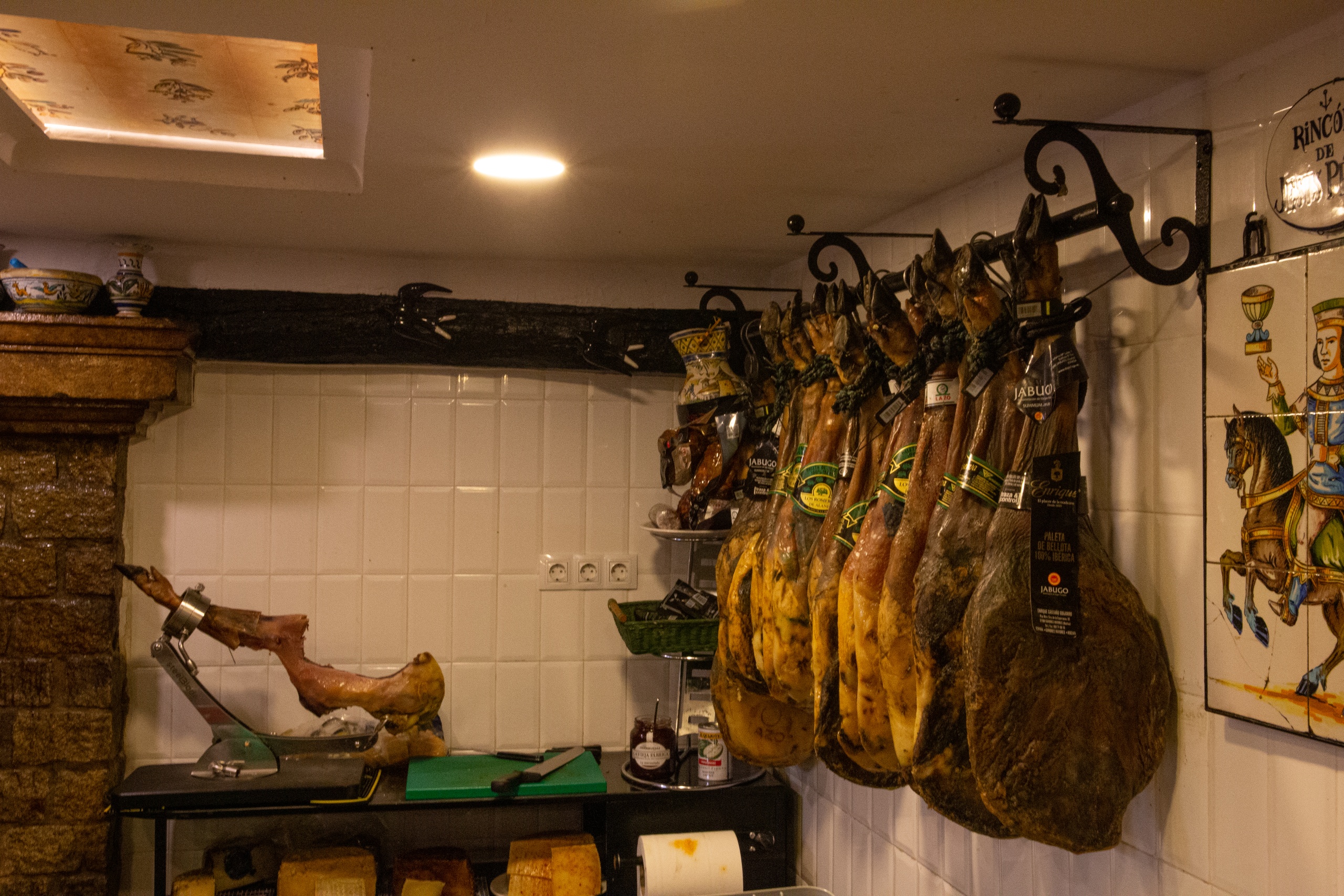 Cured hams hanging in a tiled room with a cutting station and jamón holder.