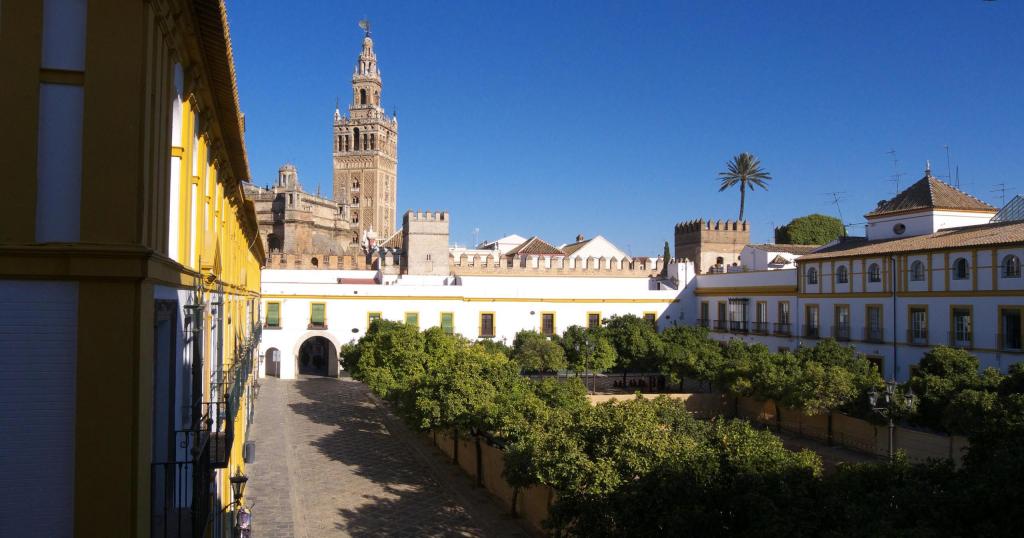 Courtyard with orange trees, yellow buildings, and a historic tower under a clear blue sky.