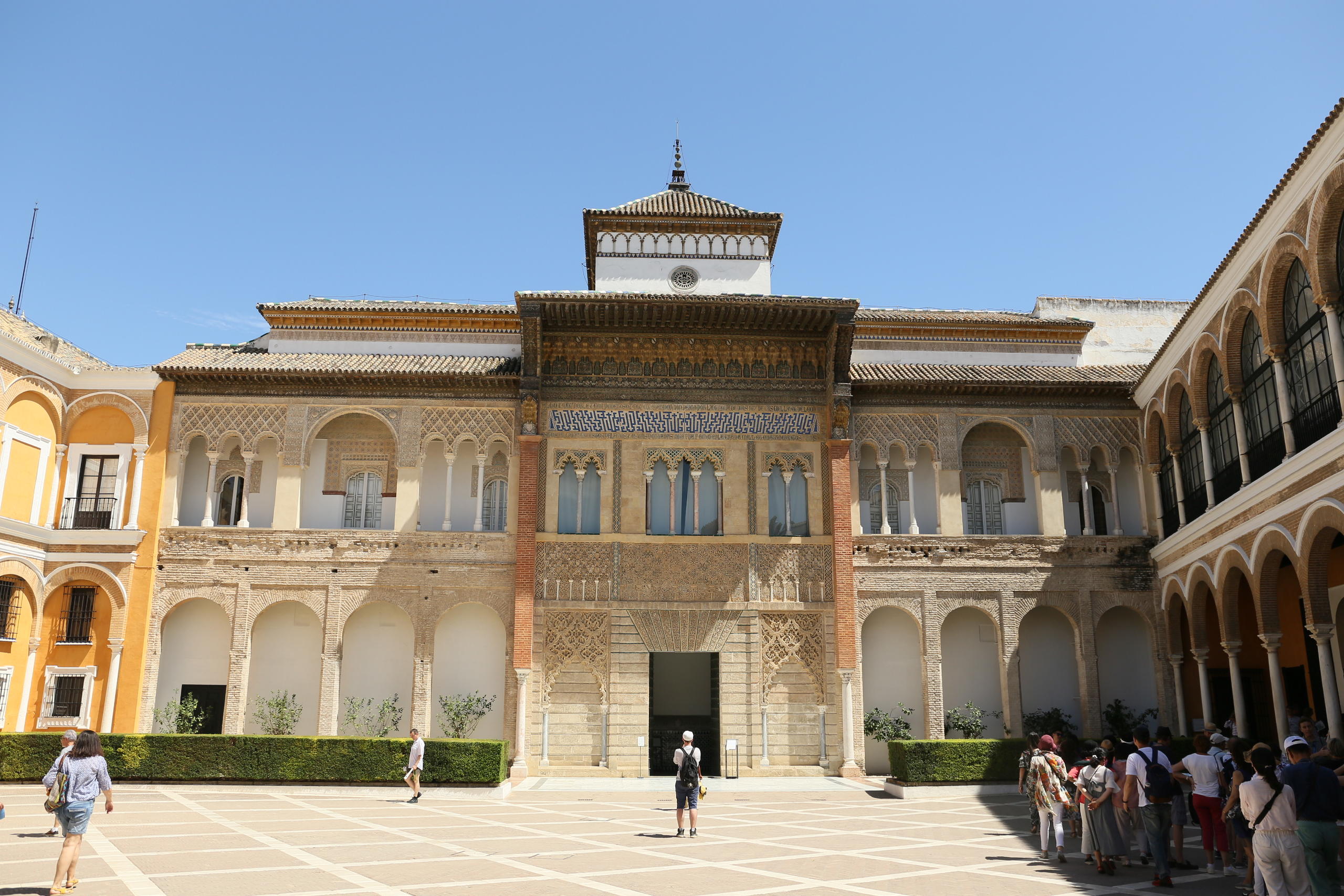 Tourists in courtyard of historic building with arched windows and detailed facade under clear blue sky.
