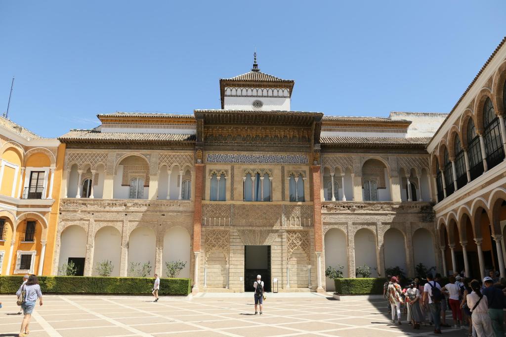 Tourists in courtyard of historic building with arched windows and detailed facade under clear blue sky.