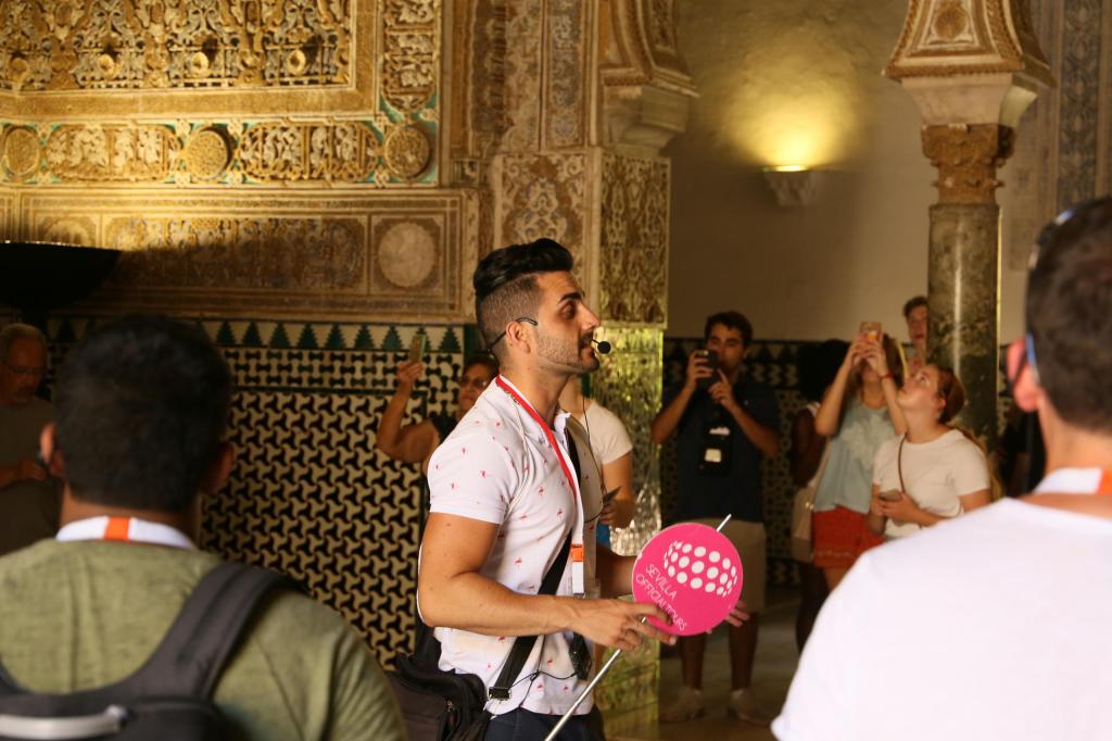 Tour guide holding a fan, speaking to a group in a decorated historic building.